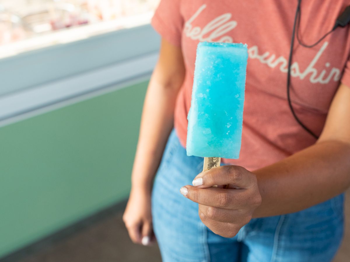 A person in a red shirt and blue jeans is holding a blue popsicle. The background shows a window with a blurry view outside.