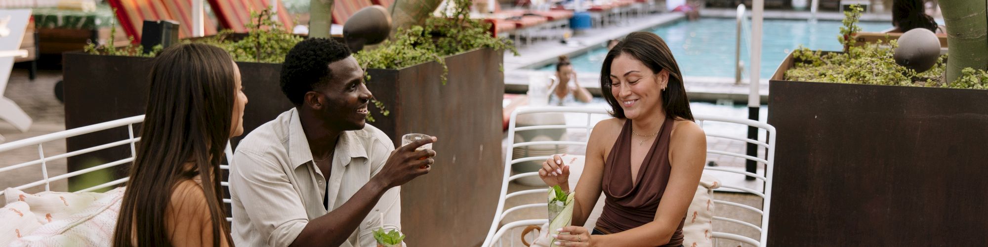Three friends chat and sip drinks on a poolside patio, lounging on chairs and a round table under leafy shade.