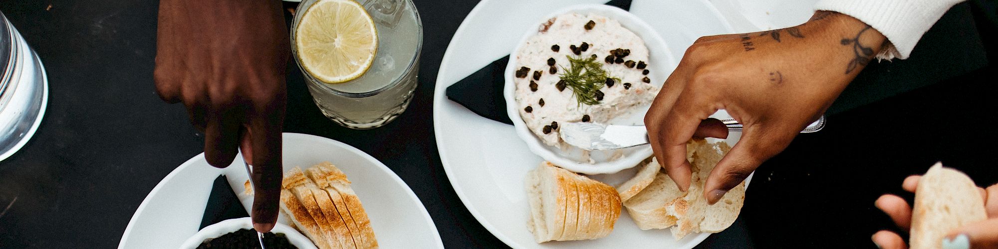 Two people enjoying a meal with bread, dips, and a drink with a slice of lemon, sharing food across a table.