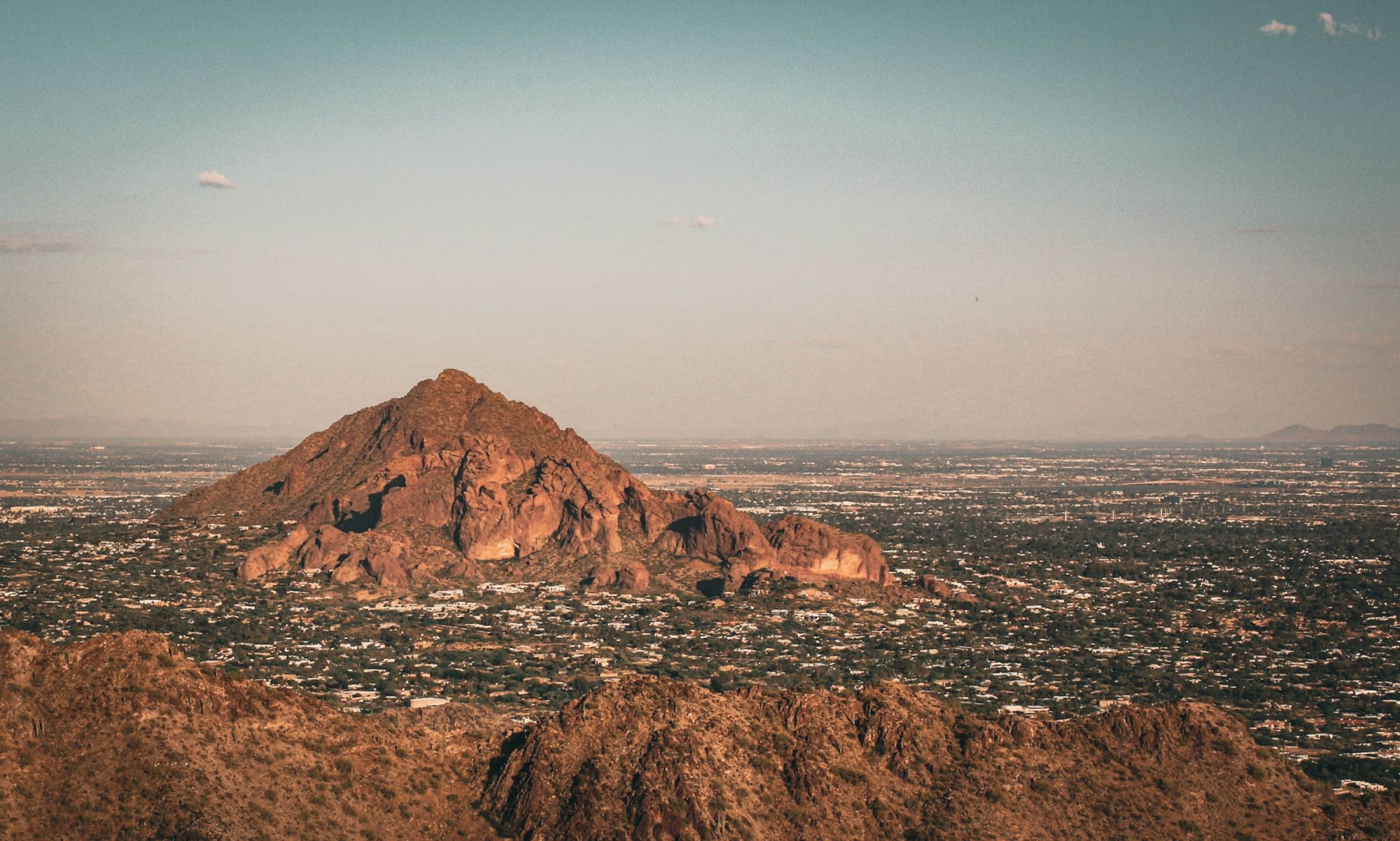 The image shows a mountainous landscape with a prominent rocky mountain in the background, surrounded by a sprawling urban area under a clear sky.