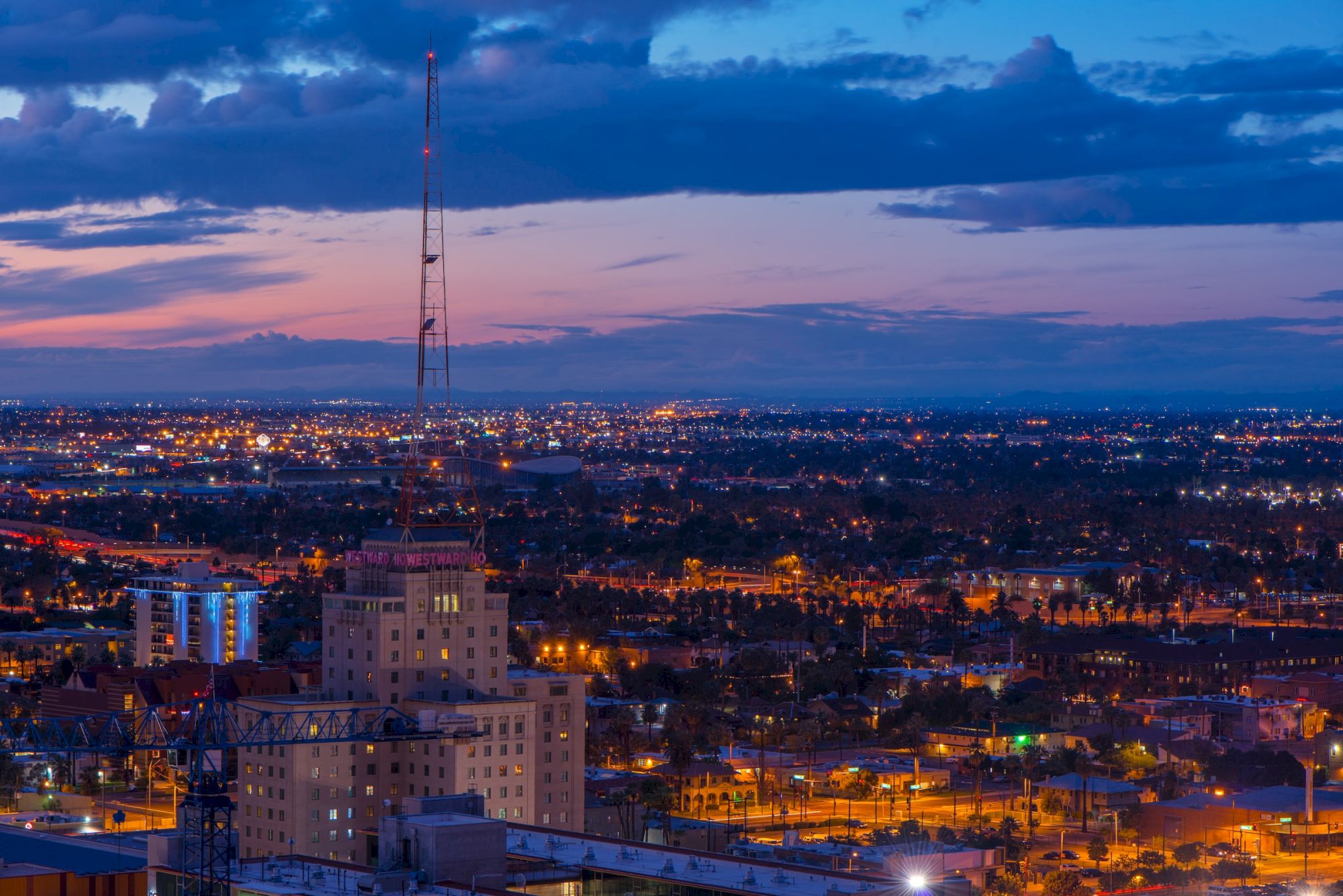 The image depicts a cityscape at dusk with illuminated buildings, streets, and a transmission tower, under a sky transitioning from sunset to night.