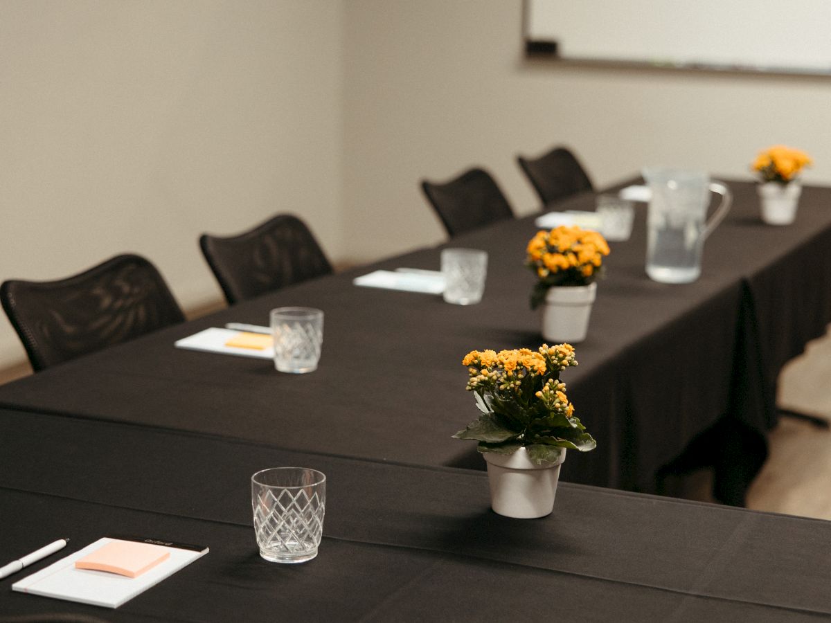 A conference room setup with a long table, black cloth, chairs, notepads, pens, glasses, flowers, and a whiteboard on the wall.