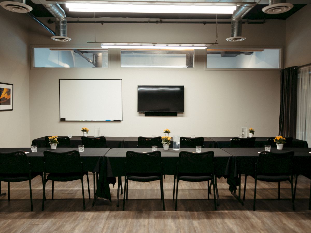 A conference room with arranged tables, chairs, a whiteboard, a TV, and potted plants on the tables.