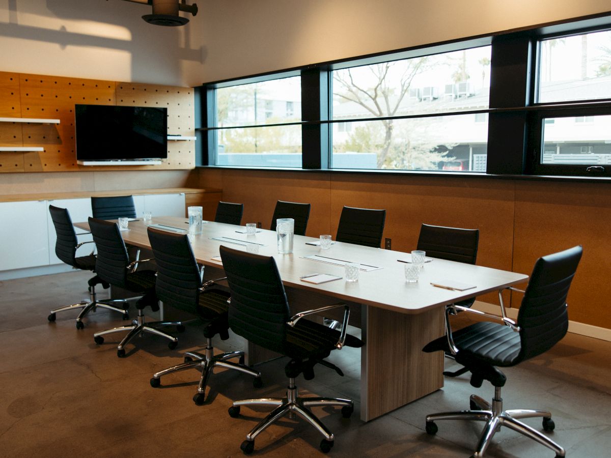 A modern conference room with a long table, black chairs, glassware, and a wall-mounted TV.