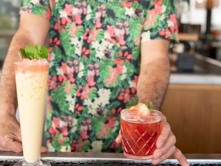 A person in a floral shirt is holding two drinks at a bar: a tall, creamy beverage in a glass and a shorter red drink garnished with fruit.