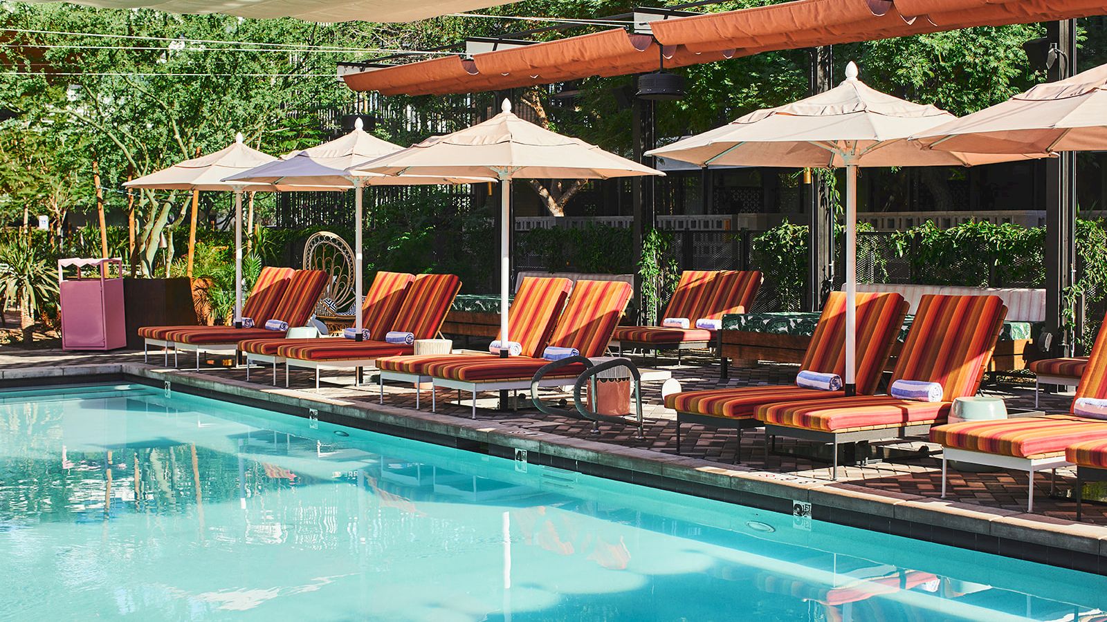 This image shows a swimming pool area with striped lounge chairs, umbrellas, and various plants in the background, creating a relaxing atmosphere.