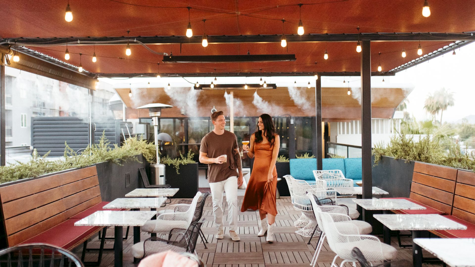 An outdoor patio restaurant with string lights, where a man and woman stand together holding drinks.