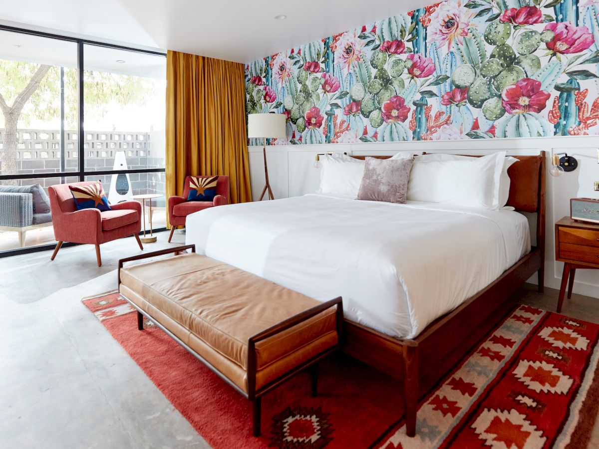 A stylish bedroom with a floral accent wall, king-size bed, leather bench, vibrant rug, and seating area with modern chairs and a patio view through glass doors.