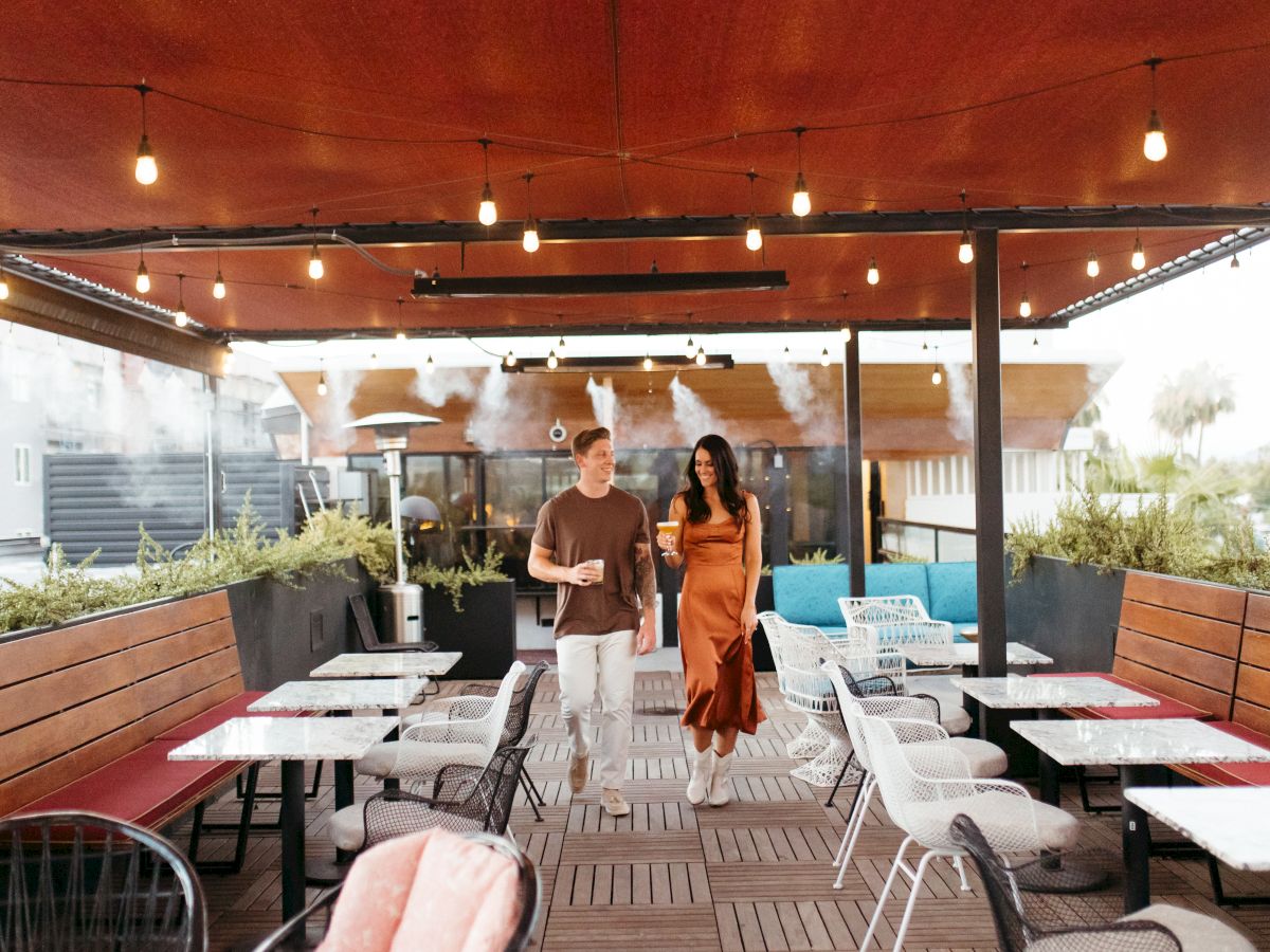 Two people walk in an outdoor restaurant with hanging lights, wooden ceiling, tables, and chairs around them. They appear to be enjoying their time.