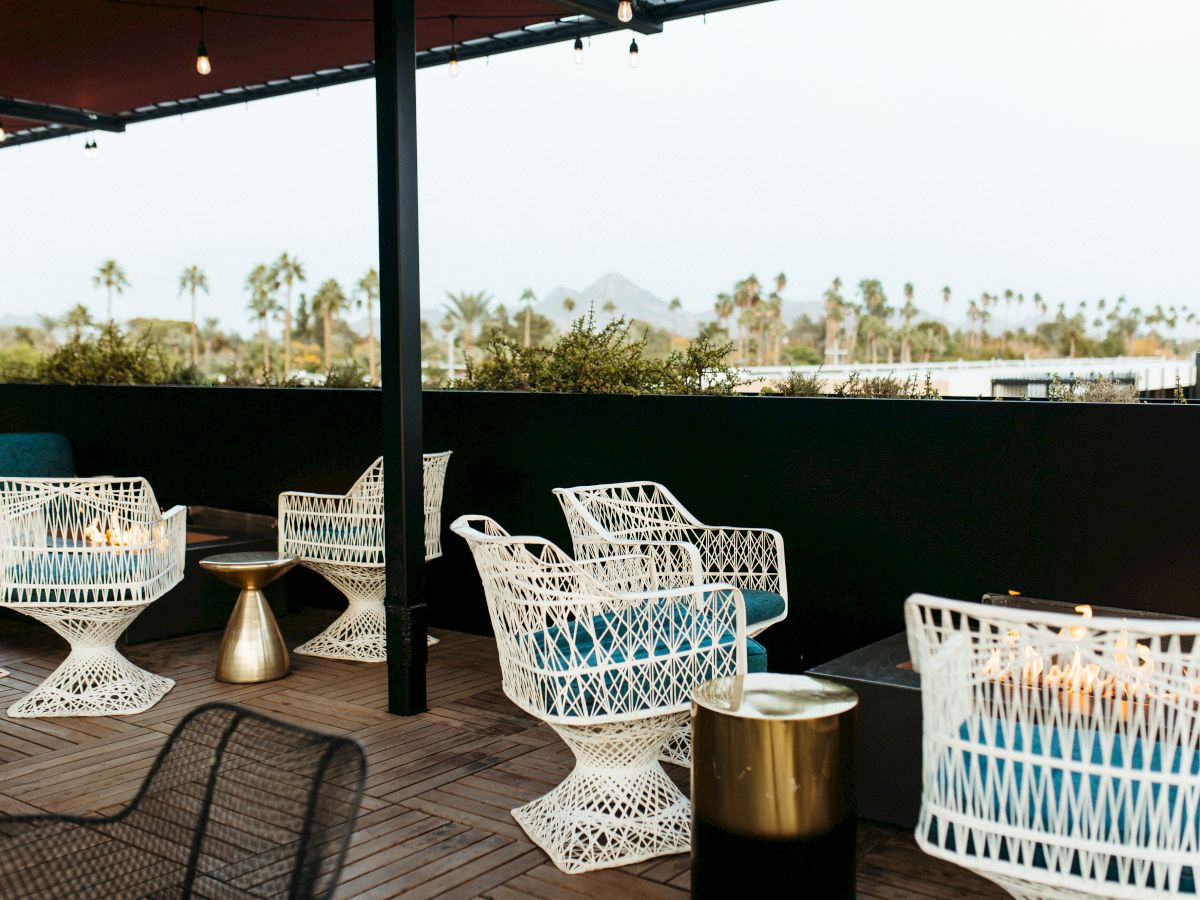 A stylish outdoor patio with modern white chairs, small side tables, and fire pits, overlooking a scenic view with palm trees in the background.