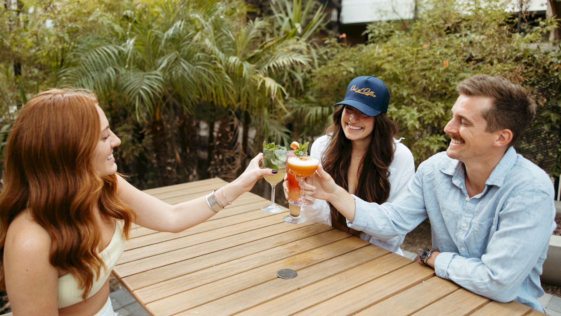 Three friends clink beer glasses at a sunlit outdoor table, sharing a toast and smiles.