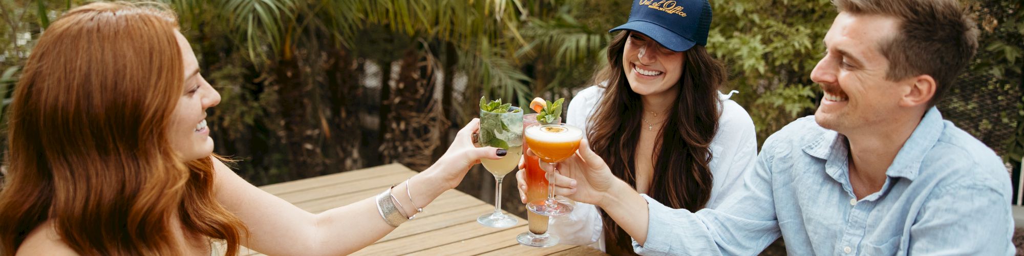 Three friends clink beer glasses at a sunlit outdoor table, sharing a toast and smiles.
