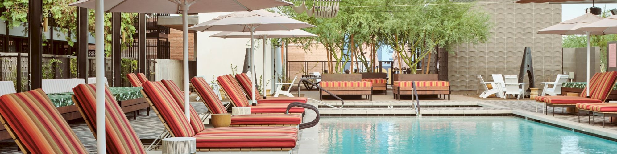 The image shows a swimming pool area with red-striped lounge chairs, umbrellas, and nearby greenery under a blue sky.