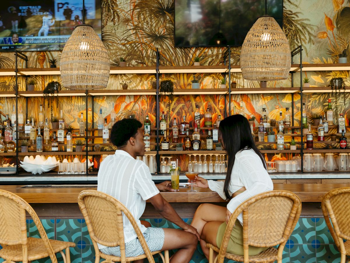 A couple sits at a bar with tropical décor, rattan chairs, hanging lights, and a wall lined with various bottles and two TVs.