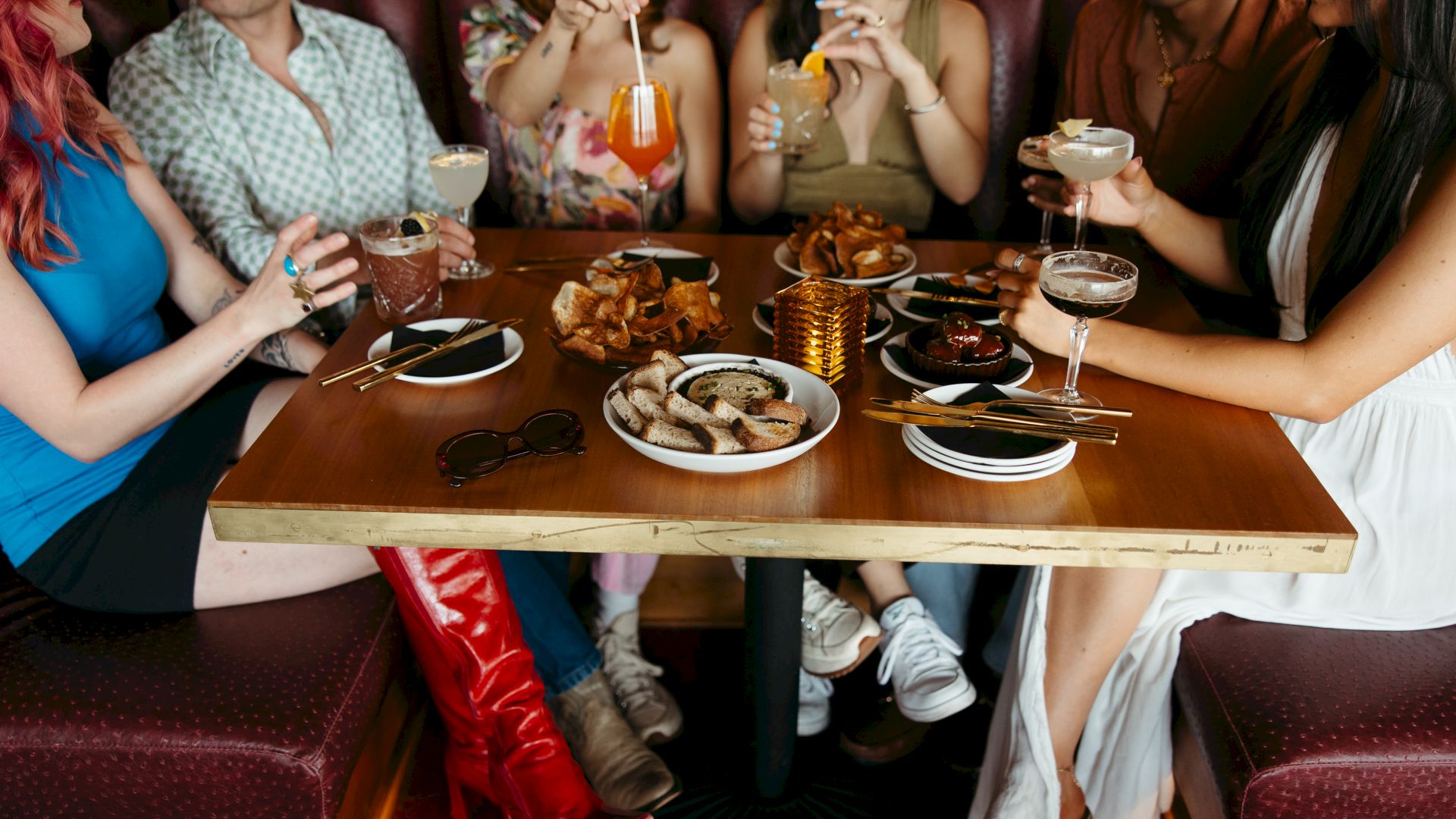 A group of friends sits around a table enjoying drinks and plates of fried finger foods, legs under the table, cozy dining vibe.