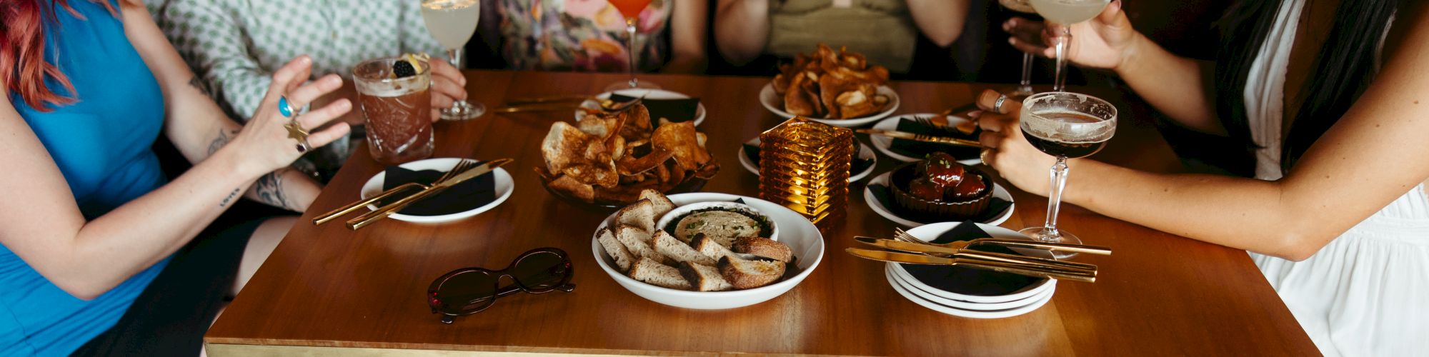 A group of friends sits around a table enjoying drinks and plates of fried finger foods, legs under the table, cozy dining vibe.