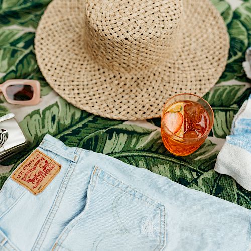 A straw hat, sunglasses, a camera, light blue denim shorts, a striped towel, and a drink with an orange slice, all on a leafy backdrop, are shown.