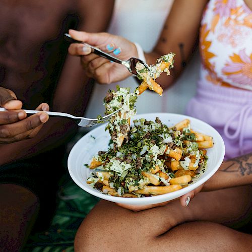 Two people are sharing a dish of loaded fries topped with greens and sauce. One person is holding a fork with fries, while the other holds the bowl.