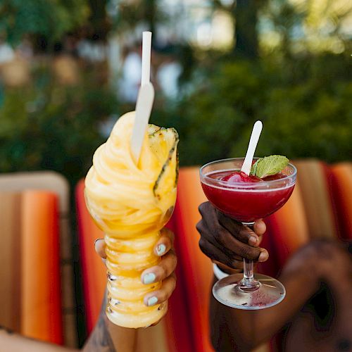 Two hands holding fruity drinks with straws, one appears to be an orange smoothie, and the other a red cocktail with a lime slice.