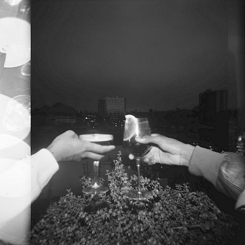 Two hands are clinking glasses outdoors at night, against the backdrop of a city skyline with buildings and lights in the distance.