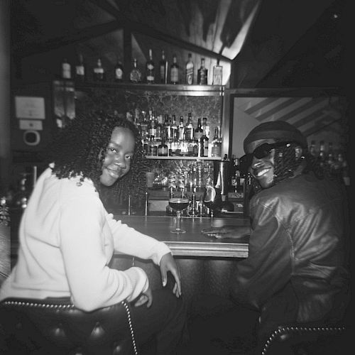 Two people are seated at a bar, smiling and facing the camera. Various bottles of alcohol are visible on shelves behind the bar.