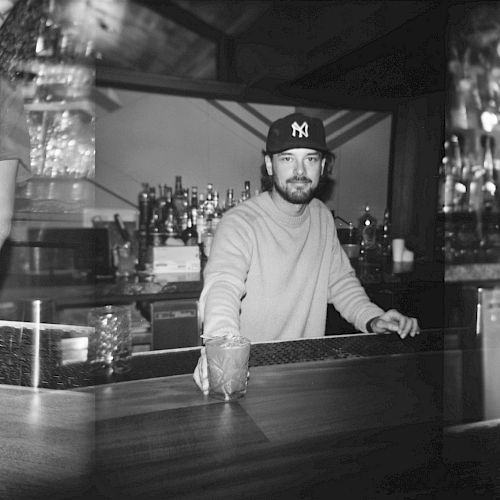 A man in a cap stands behind a bar, holding a glass, with liquor bottles and barware visible in the background.