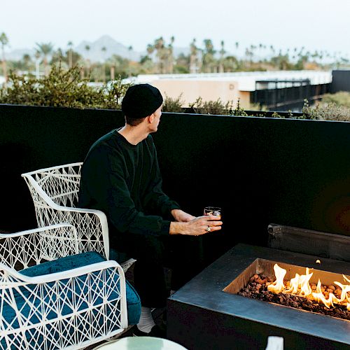 A person sits by a fire pit on a patio, looking out at a scenic view with mountains and palm trees in the distance, holding a drink.