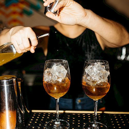 A bartender is preparing cocktails, pouring liquid into two wine glasses filled with ice. The focus is on the drinks and the bartender's hands.