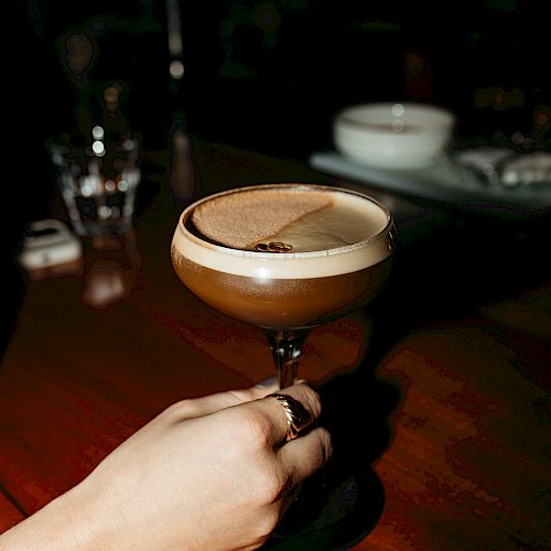 A hand holding a cocktail glass with a brown drink on a wooden surface, with glassware and a bowl visible in the background.