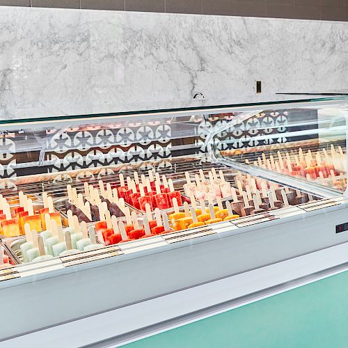 A display counter filled with various colored popsicles inside an ice cream shop, with a sign board listing different flavors on the wall behind it.