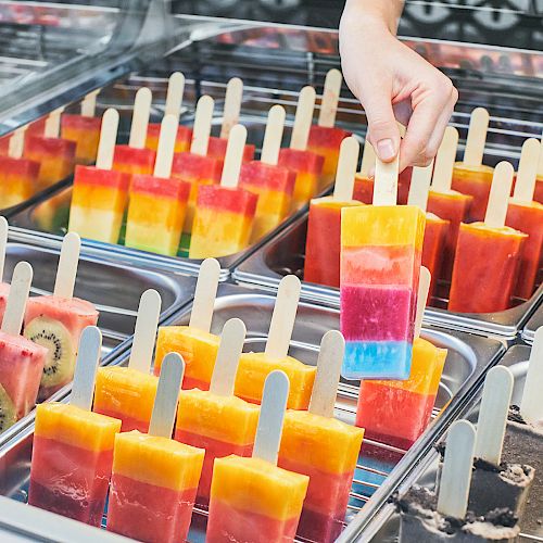 A hand picks up a colorful layered popsicle from a display case filled with various vibrant and fruity popsicles arranged in trays.