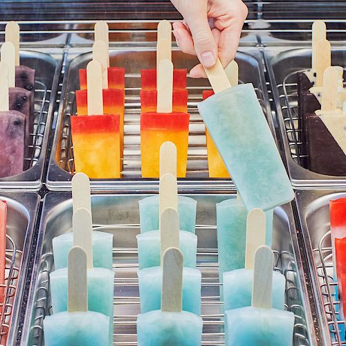 The image shows an assortment of colorful popsicles displayed in metal trays, with a hand picking one up.