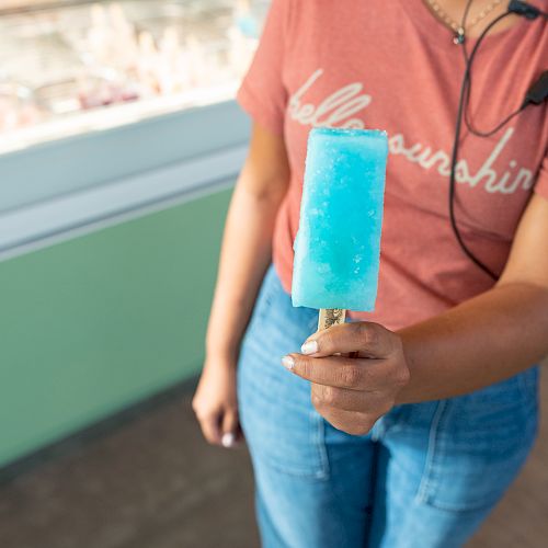 A person wearing a reddish t-shirt holds a blue popsicle in their hand while standing indoors near a window with blurred background.