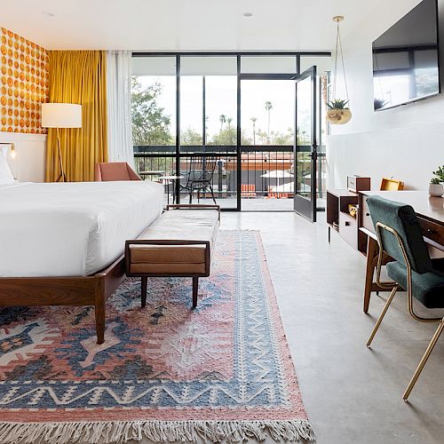 A modern bedroom with a large bed, a patterned rug, a desk, a TV, and a balcony view. The room features colorful, geometric wallpaper.