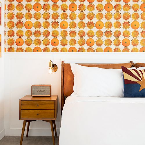 A stylish bedroom with vibrant orange wallpaper, a mid-century nightstand, a wall-mounted light, and a neatly made bed with a decorative pillow.