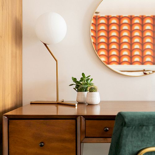 A retro-styled desk with a lamp, potted plants, and a green chair is in front of a round mirror reflecting a patterned window curtain, in a cozy setup.