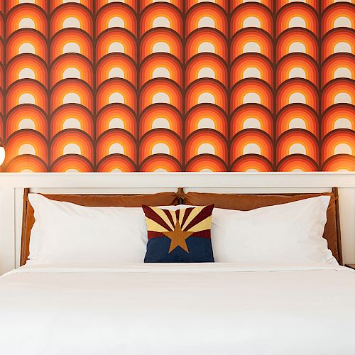 A neatly made bed with a geometric orange and red patterned wall, flanked by two pendant lights and two nightstands holding a speaker and plant.