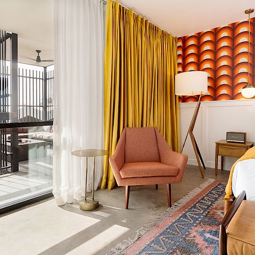 A stylish room with a retro design featuring a patterned orange wall, yellow curtains, an armchair, a bed with white sheets, and a balcony.