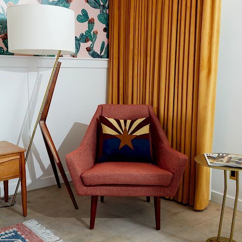 A cozy corner with an armchair, cactus wallpaper, gold curtains, a floor lamp, and a side table with books and a candle.