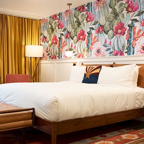 A cozy bedroom featuring a floral wallpaper, white linens, a wooden bed frame, a floor lamp, and a patterned rug.