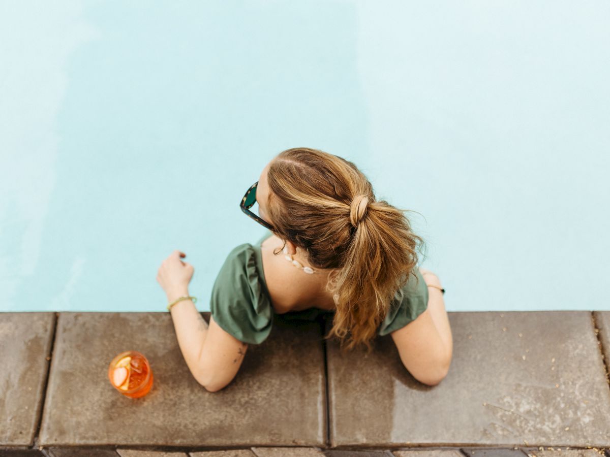 A person with a ponytail is sitting by a pool, resting on the edge, with an orange drink placed beside them on the pool deck.