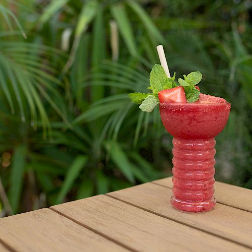 A red smoothie or drink garnished with mint leaves and strawberry slices in a unique glass, placed on a wooden table with greenery in the background.