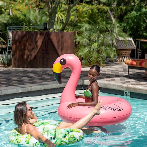 Two people are in a pool, one on a pink flamingo float and the other on a green float. Surrounded by lush greenery, they seem to be enjoying.