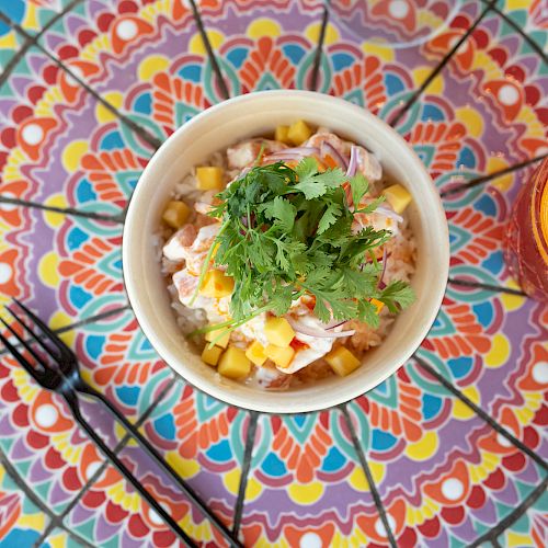 A colorful dish with cilantro, mango, and shrimp in a bowl, with a glass of fruity drink; all on a vibrant, patterned table.