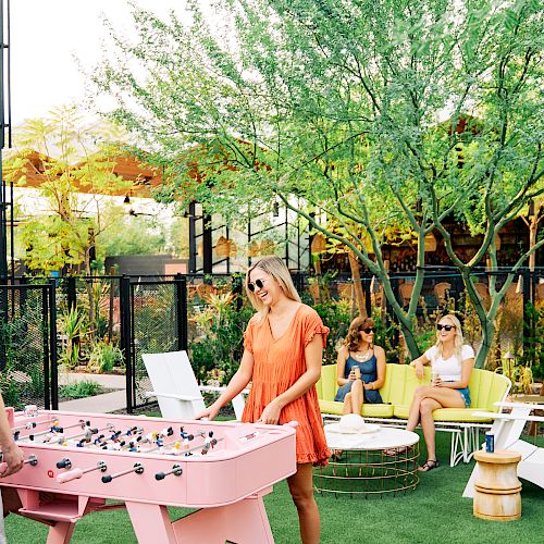 People enjoying an outdoor area with a pink foosball table, lounging on bright furniture, and surrounded by greenery and modern decor.