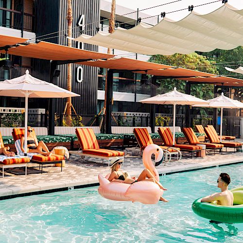 People are lounging by the pool on striped chairs with umbrellas, while others are floating on inflatable toys in the water.