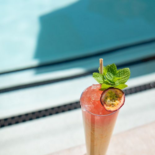 A tall glass of a colorful drink garnished with a passion fruit slice and mint leaves, placed near a poolside on a sunny day.