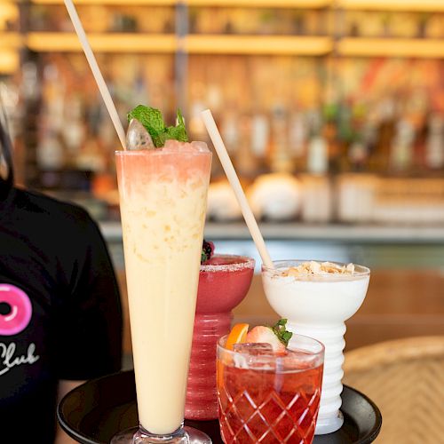 A hand holds a black tray with four drinks: a tall yellow smoothie, a pink beverage, an ice-filled red drink with lemon garnish, and a white bowl with food.