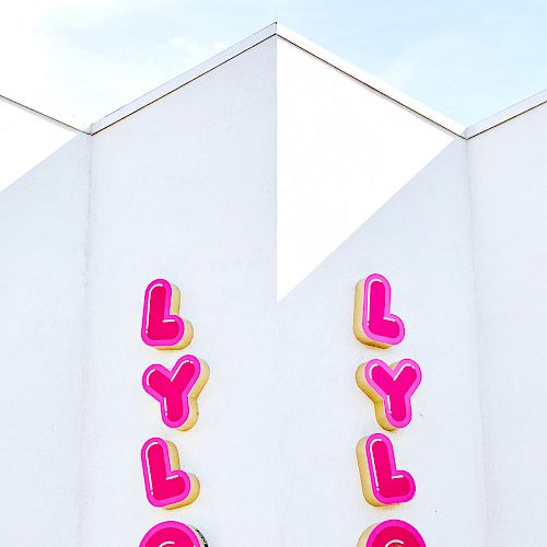 The image shows a white building with two vertical signs displaying the word "LYLO" in bright pink and yellow letters, under a blue sky.