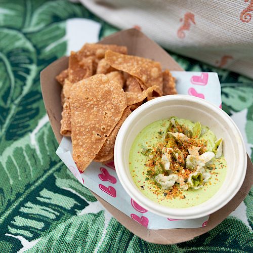 A bowl of dip sits alongside a serving of crispy chips on a paper tray with decorative paper, placed on a green leafy-patterned fabric.